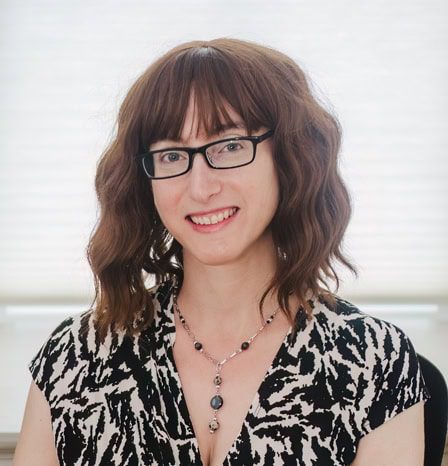 Cute woman with wavy brown hair, black glasses, and a patterned black and white top on a bright, airy background.