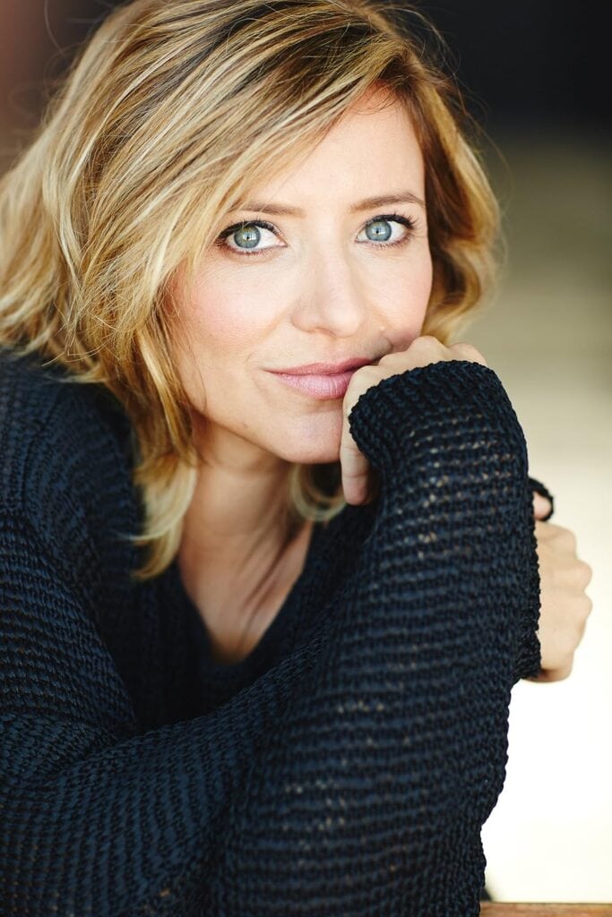 Blonde woman with blue eyes wearing a black knit sweater, close-up portrait, casual and cozy fashion, natural beauty, confident expression, friendly vibe, outdoor setting, sunny weather.