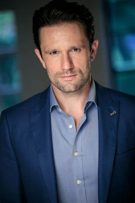 A man in a navy blue suit and light blue shirt, professional headshot, confident expression, modern office background.